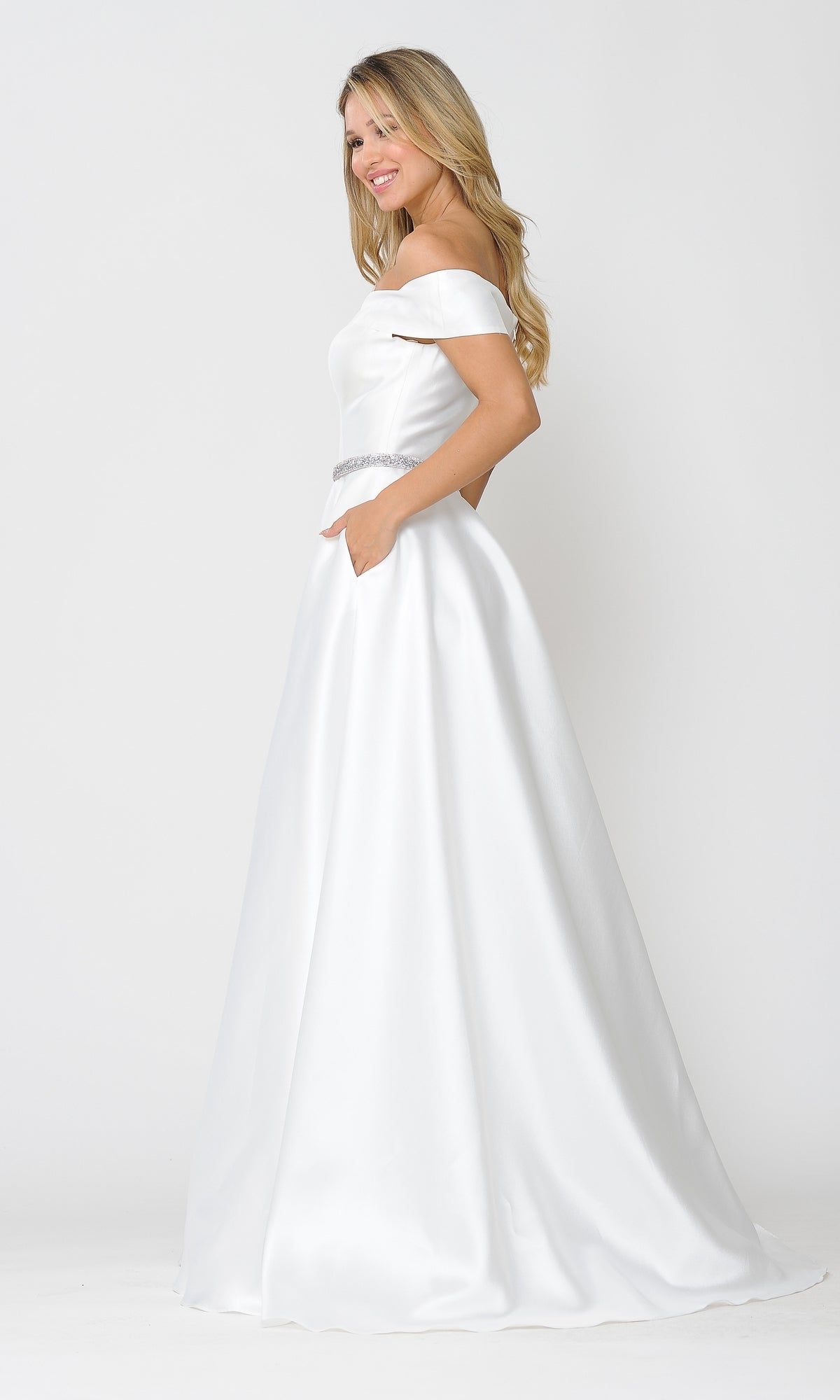 A woman stands against a white background wearing the Plunging V-Neck Long Mikado Prom Ball Gown 8680, featuring an off-the-shoulder design, full skirt, silver belt detail, and hands in pockets.